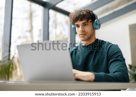 E-learning concept. Focused european guy with wireless headphones looking at laptop, watching online tutorial, lecture or class sitting in university classrom or library