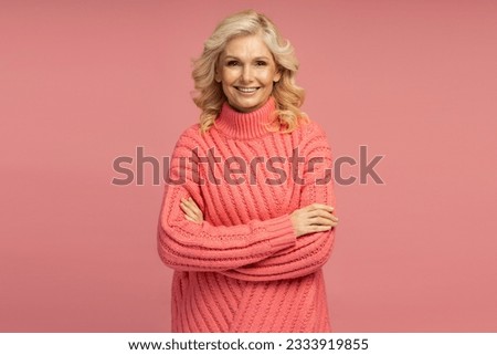Beautiful mature blonde woman wearing pink stylish pullover, crossed arms, smiling, looking at camera, standing sideways isolated on pink background. Store, shopping, sale concept 