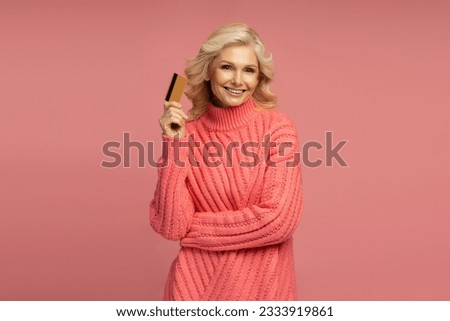 Blonde mature woman wearing stylish winter pink sweater, holding golden credit card, smiling a beautiful toothy smile at camera, isolated on pink coloured background