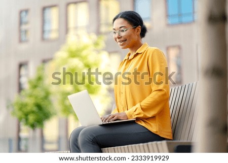 Beautiful smiling African American woman, worker holding laptop computer, having video call sitting on bench outdoors. Modern technology, video conference, online lesson concept 