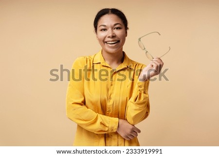 Smiling African American woman looking at camera with eyeglasses in hand isolated on beige background, copy space. Successful business, career concept