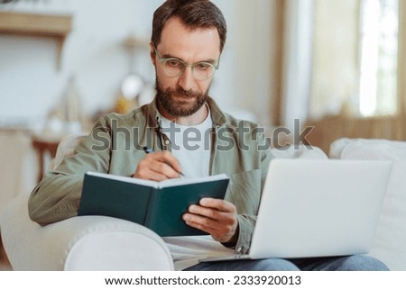 Handsome smiling bearded businessman wearing eyeglasses taking notes, studying at home. Smart student learning language, online education concept 