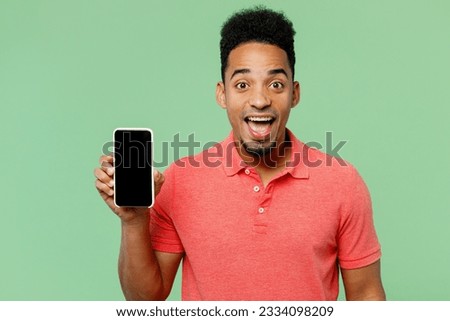 Young smiling happy man of African American ethnicity he wears pink t-shirt hold in hand use mobile cell phone with blank screen workspace area isolated on plain pastel light green background studio