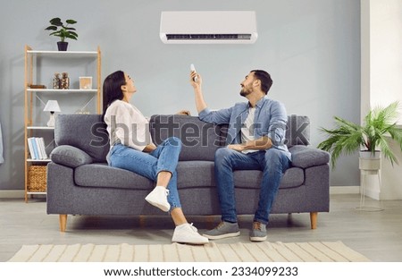 Young happy man and woman turning on air conditioner sitting on sofa at home. Smiling couple of homeowners enjoying cool conditioned air using remote resting on couch together in living room. Royalty-Free Stock Photo #2334099233