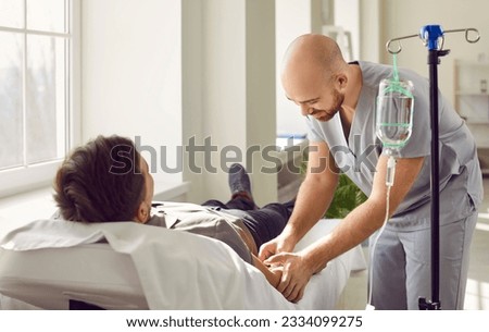 Nursing staff at clinic or hospital gives intravenous vitamin drip or medicine infusion to patient. Nurse personnel inserts venous IV line needle in arm vein of adult man lying on bed in medical ward Royalty-Free Stock Photo #2334099275
