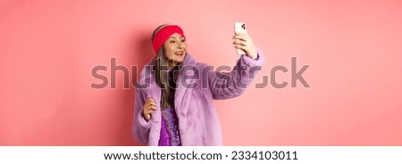 Fashion concept. Beautiful asian senior lady taking selfie on smartphone, posing in party glittering dress and purple faux coat, standing over pink background.