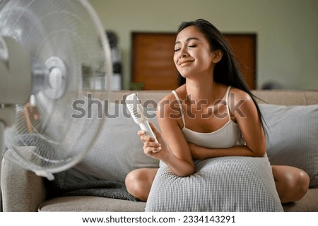A gorgeous Asian woman, feeling hot suffering from a heat attack, refreshes herself with a handy fan and an electric fan while sitting on a couch in her living room on a summer day. Royalty-Free Stock Photo #2334143291
