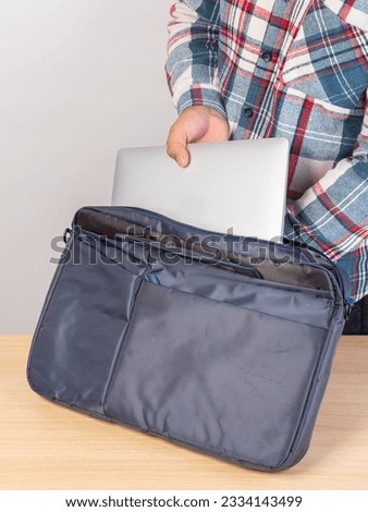 The hands of an unrecognizably cropped man in a plaid shirt put a laptop in a bag, going to work.