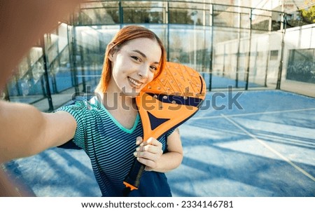 A cute paddle tennis player takes a picture of herself happily posing with a paddle tennis racket. Outdoor paddle tennis concept, women playing paddle tennis.