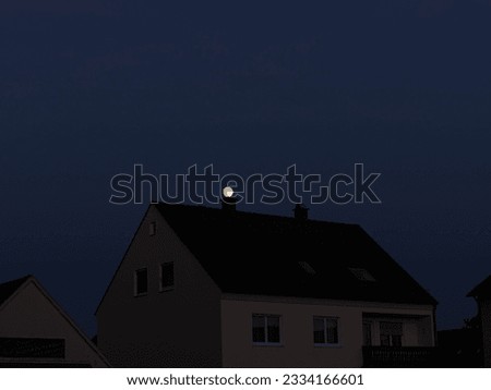 Full moon peeping from the chimney of the roof of the house. House is cream color, roof is dark tiled , sky is deep blue at dusk