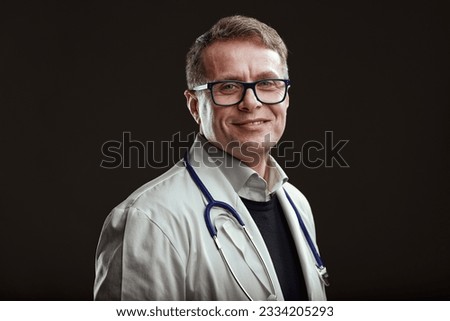 Middle-aged, bespectacled doctor smiles, passionate about his profession, gracious and content to assist people through his knowledge. Dark background portrait Royalty-Free Stock Photo #2334205293