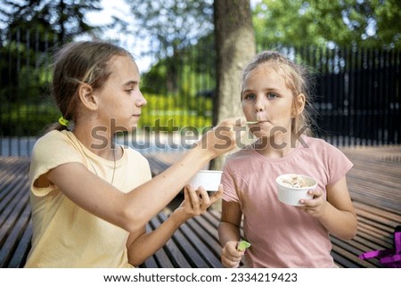 portrait of two cute blonde girls, they eat cold ice cream from paper cups. One tries a treat from another girl. A walk in the park, summer holidays and fun