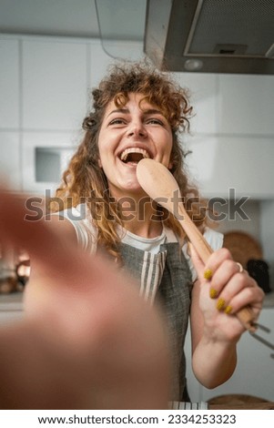 One happy young adult caucasian woman wear apron in the kitchen smile hold mixing spoon sing self portrait selfie ugc user generated content Royalty-Free Stock Photo #2334253323