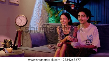 Happy Indian teenager couple sitting on cozy sofa watching favourite movie TV having fun enjoy laughing weekend together. Smilling teen boyfriend girl friend hold popcorn boul eating snack at home