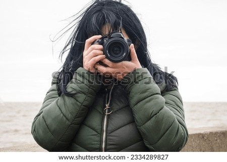 front view portrait of young tourist woman wearing green jacket, outdoors standing by the sea, with her digital camera taking a photo and looking at the camera.