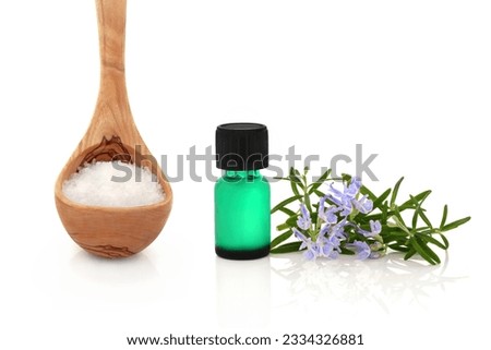 Rosemary herb leaf sprig in flower with a green aromatherapy essential oil bottle and sea salt in an olive wood ladle, over white background.