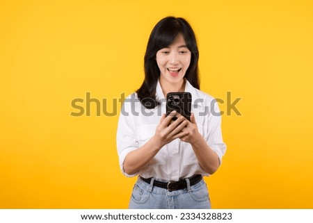 Surprised asian young woman using mobile phone with positive expression, smiles broadly, dressed in white shirt and standing isolated on yellow background. Happy adorable glad woman rejoices success. Royalty-Free Stock Photo #2334328823