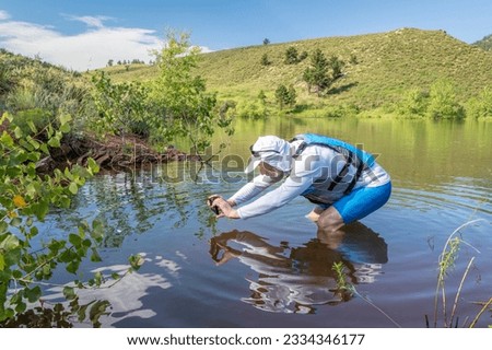 senior male paddler wearing life jacket and sun hat is shooting pictures with a phone in water near shore, Horsetooth Reservoir in Colorado