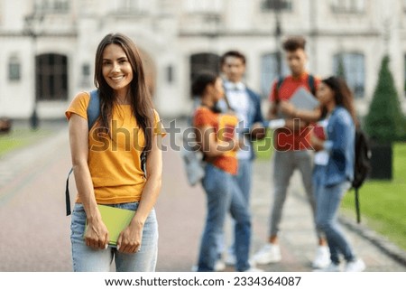 Student Life. Outdoor portrait of smiling young female posing in front her college friends at campus, beautiful millennial woman carrying backpack and workbooks, smiling at camera, selective focus