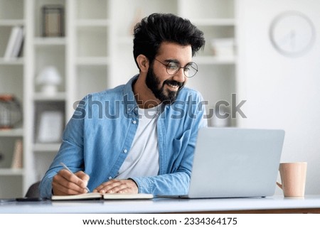 Online Lesson. Smiling Young Indian Man Using Laptop And Taking Notes While Sitting At Desk At Home, Smiling Eastern Male Enjoying Remote Education And Distance Learning, Free Space Royalty-Free Stock Photo #2334364175