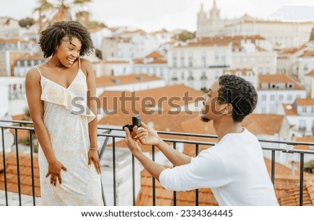 Smiling young african american guy kneeling, gives box with ring, proposes to lady in dress in city, enjoy date, travel, outdoor. Love, relationships, vacations and romance, marriage and engagement