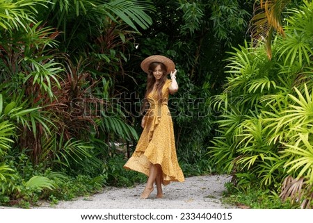 Female tourist girl with fashionable hat poses elegantly in lush green jungle. Young woman dancing charm and happiness in portrait of a tropical vacation. Royalty-Free Stock Photo #2334404015