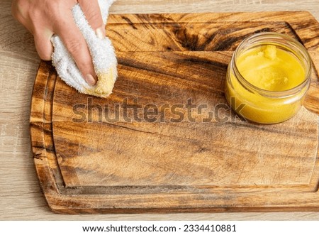 Woman hands apply homemade beeswax wood treatment polish to restore natural wood cutting board. Beeswax, olive oil and essential oil, soft cloth and mixture in glass jar. Polishing wood.  Royalty-Free Stock Photo #2334410881