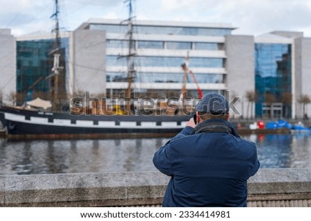 Man wearing a flat cap and blue jacket takes a picture with his smartphone of a sailing ship across the riverbank of the Liffey in Dublin, Ireland