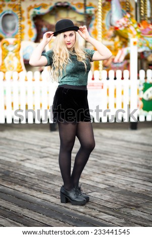 Portrait of a beautiful confident blond blue eyed teenage girl with long hair wearing a bowler hat, glittery t-shirt dark red lipstick standing alone in front of a fairground ride with a white fence.