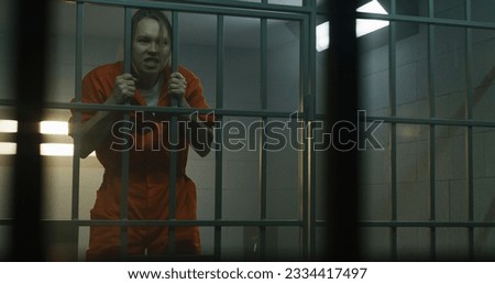 Insane female prisoner in orange uniform yells at neighbor inmate, jumps on bars in prison cell. Woman criminal serves imprisonment term for crime in jail. Detention center. View through metal bars. Royalty-Free Stock Photo #2334417497