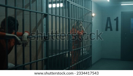 One female prisoner in orange uniform stands behind metal bars, another sits on the bed in prison cell. Women serve imprisonment terms for crimes in jail. Depressed inmates in detention center. Royalty-Free Stock Photo #2334417543