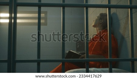 Female prisoner in orange uniform sits on bed behind metal bars, reads Bible in prison cell, looks at barred window. Woman criminal serves imprisonment term for crime in jail or correctional facility. Royalty-Free Stock Photo #2334417629