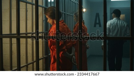 Two female prisoners, inmates in orange uniforms stand facing the metal bars in front of prison cells. Prison officer walks, watches women criminals in jail. Detention center or correctional facility. Royalty-Free Stock Photo #2334417667