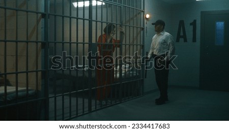 Jailer with police baton stands near prison cell, talks to female prisoner in orange uniform through metal bars. Women serve imprisonment terms in jail. Detention center or correctional facility. Royalty-Free Stock Photo #2334417683