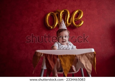 Portrait of Little boy of one year in festive hat sits in childrens chair with fabric flags. Kid on red isolated background with foil gold balloons One, on textured wall celebrating holiday at home