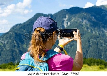 During hiking a woman takes a picture of landscapes in mountains using a smartphone. Close back view of women in pink t-shirt with trekking rucksack taking pictures with smartphone in mountains.