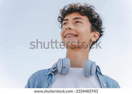 Portrait of smiling curly haired teenager with braces wearing headphones looking away standing on the street. Inspiration, technology concept Royalty-Free Stock Photo #2334483743