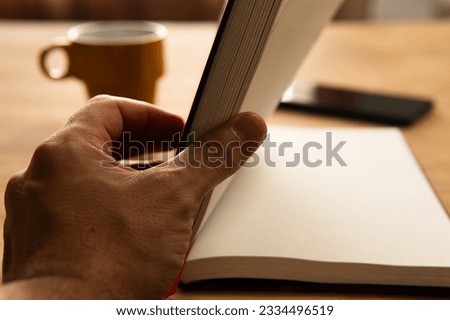 Hand opening a book with a cup of tea and mobile phone next to it on wooden table