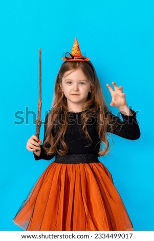 Halloween Party girl. Funny child girl in witch costume celebrates Halloween with magic wand and pumpkin pot on bright blue background