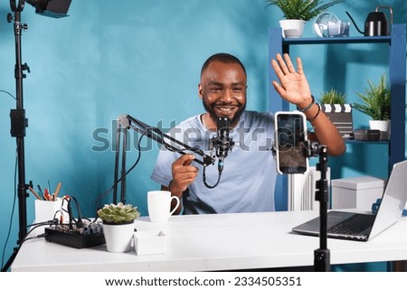 Smiling blogger waving hi and recording video on smartphone in home studio. Cheerful young african american vlogger streaming on social media using mobile phone on tripod