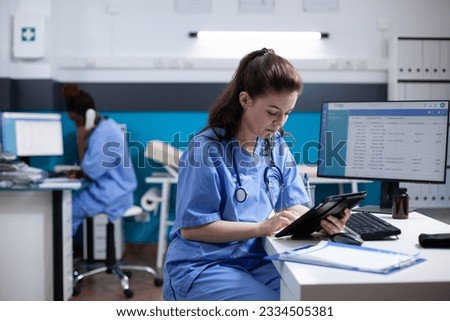 Young nurse checking appointments list on digital tablet in busy medical office. Adult woman healthcare specialist working with technology at hospital desk with stethoscope around neck