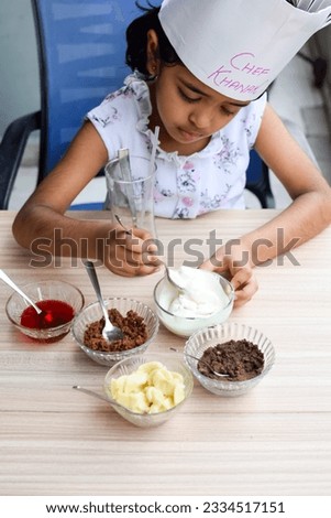 Cute Indian chef girl preparing sundae dish as a part of non fire cooking which includes vanilla ice cream, brownie, coco powder, freshly chopped fruits and strawberry syrup. Little kid preparing food
