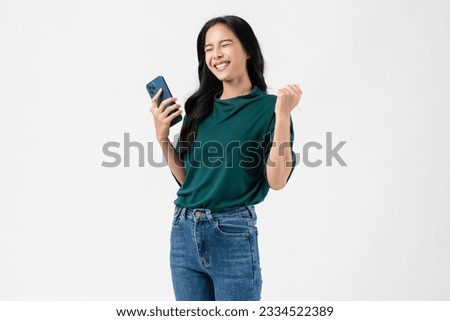 Cheerful young Asian woman holding smartphone with fists clenched celebrating victory expressing success on white background. Royalty-Free Stock Photo #2334522389