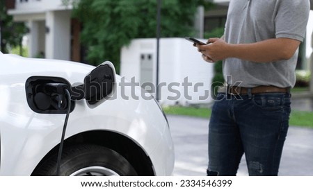 Electric car plug into home charging station to recharge battery with electricity by EV charger, man using smartphone in background. Future lifestyle of sustainable clean energy utilization. Peruse