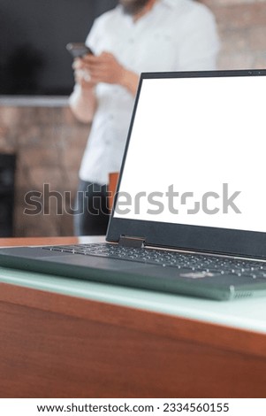 A laptop with a blank screen on a work desk with an unrecognizable person out of focus in the background who uses a smartphone to chat. Home office space.