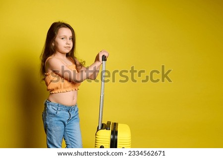 Beautiful little child girl with long hair, dressed in blue jeans and orange top, posing with suitcase, looking at camera, isolated on yellow studio background. Trip. Travel. Tourism. Weekend getaway