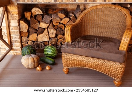 A comfortable rattan wicker chair sits on the veranda. Stacked firewood in a niche at the back. Nearby are vegetables and watermelons

