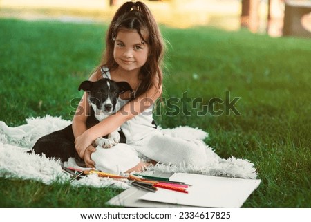A beautiful, sweet little girl is spending time with her pet dog and drawing a picture with colored pencils in the park on the green grass. A happy childhood. Drawing