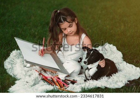 A beautiful, sweet little girl is spending time with her pet dog and drawing a picture with colored pencils in the park on the green grass. A happy childhood. Drawing