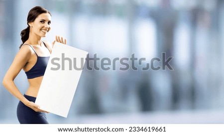 Photo of happy smiling beautiful young brunette woman in fitness wear showing blank paper signboard with copy space area. Fit girl holding mock up placard, over blurred modern gym center background.